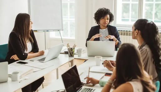 Réunion d’équipe dans une salle de travail lumineuse, avec quatre personnes discutant autour d’une table, ordinateurs portables et documents devant elles